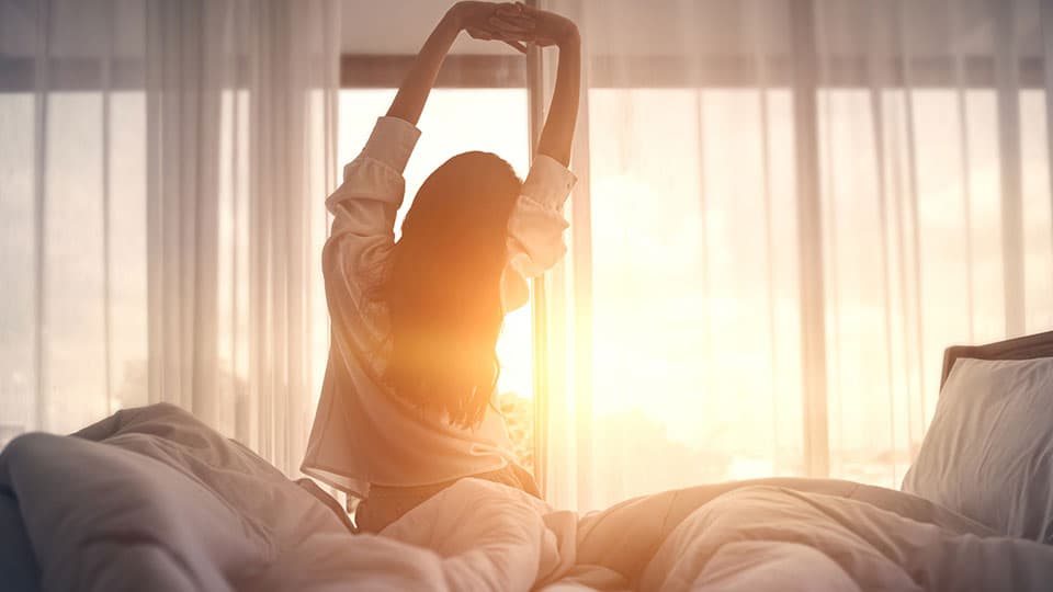 A woman sat in front of a window with the sun brightly rising, stretching in bed.
